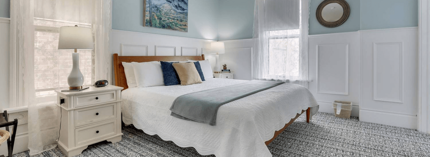 Cozy bedroom featuring a bed with decorative pillows, a nightstand, and natural light from large windows.