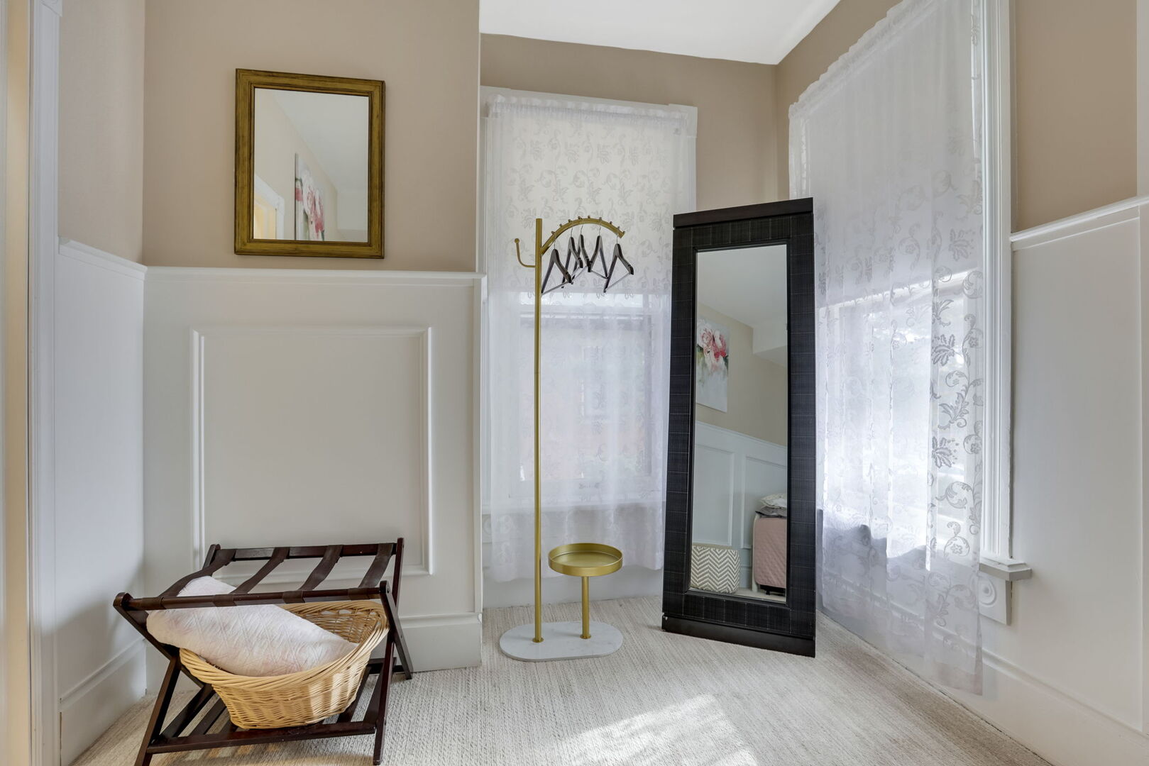 A cozy dressing area with neutral walls, white wainscoting, a gold-framed mirror, a full-length mirror, and a luggage rack.