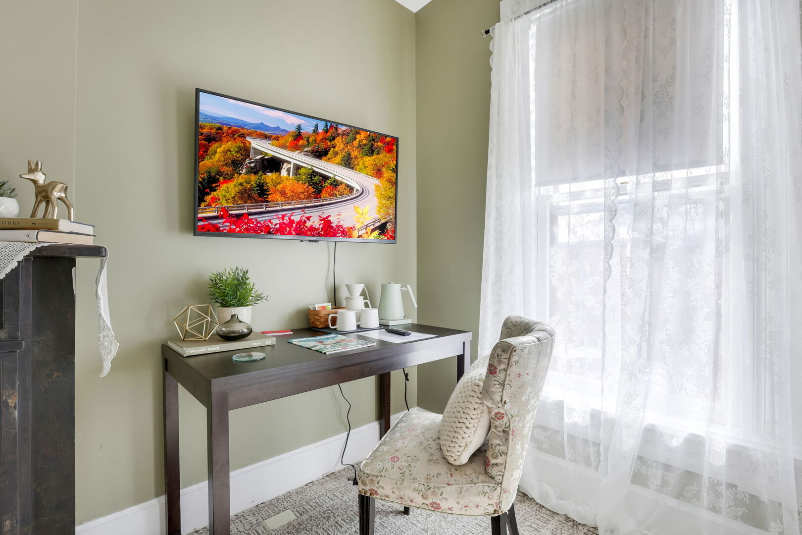 A cozy corner with olive green walls, featuring a wall-mounted TV, a dark wooden desk with a chair, and a bright window with sheer curtains.