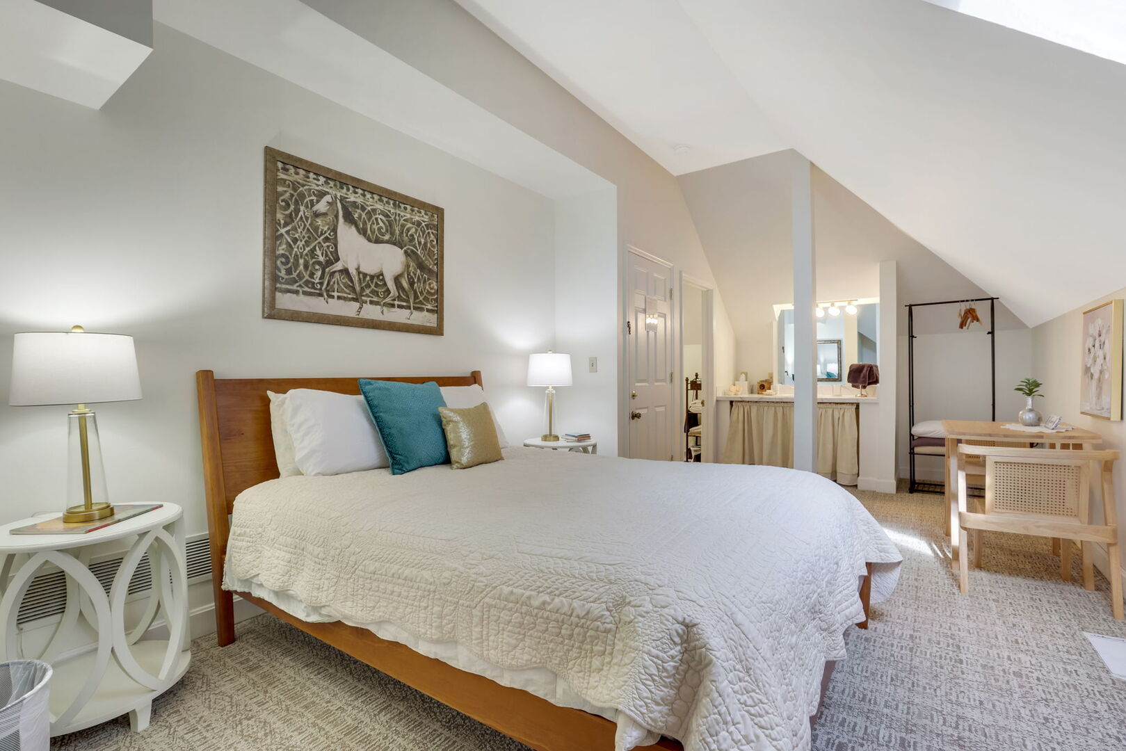 A bright bedroom with a sloped ceiling, featuring a queen bed, a framed tapestry, and a view into a separate vanity area.