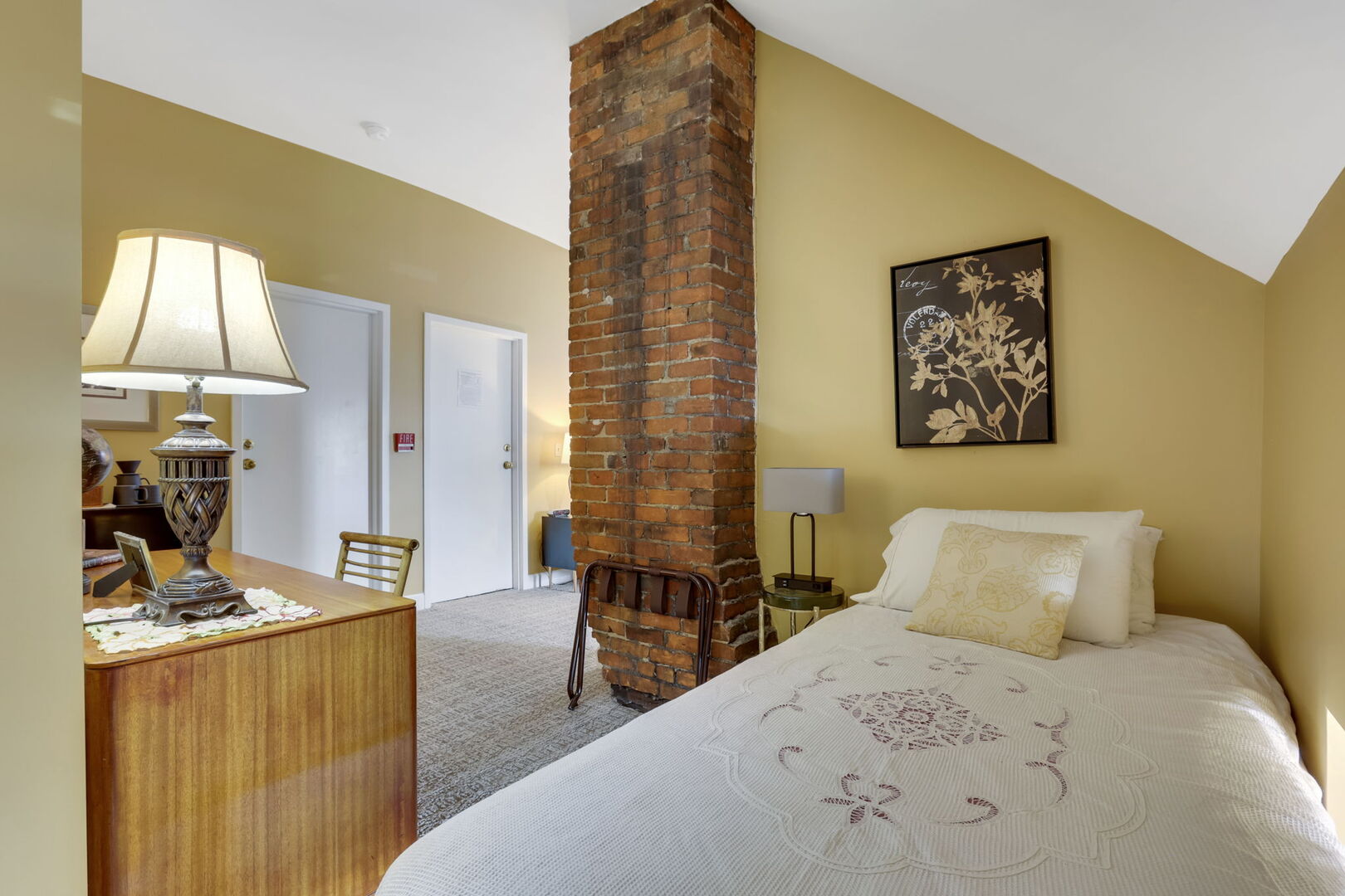 A warm third-floor bedroom with a twin bed, golden walls, a wooden beam accent, and a glimpse of a vanity area.