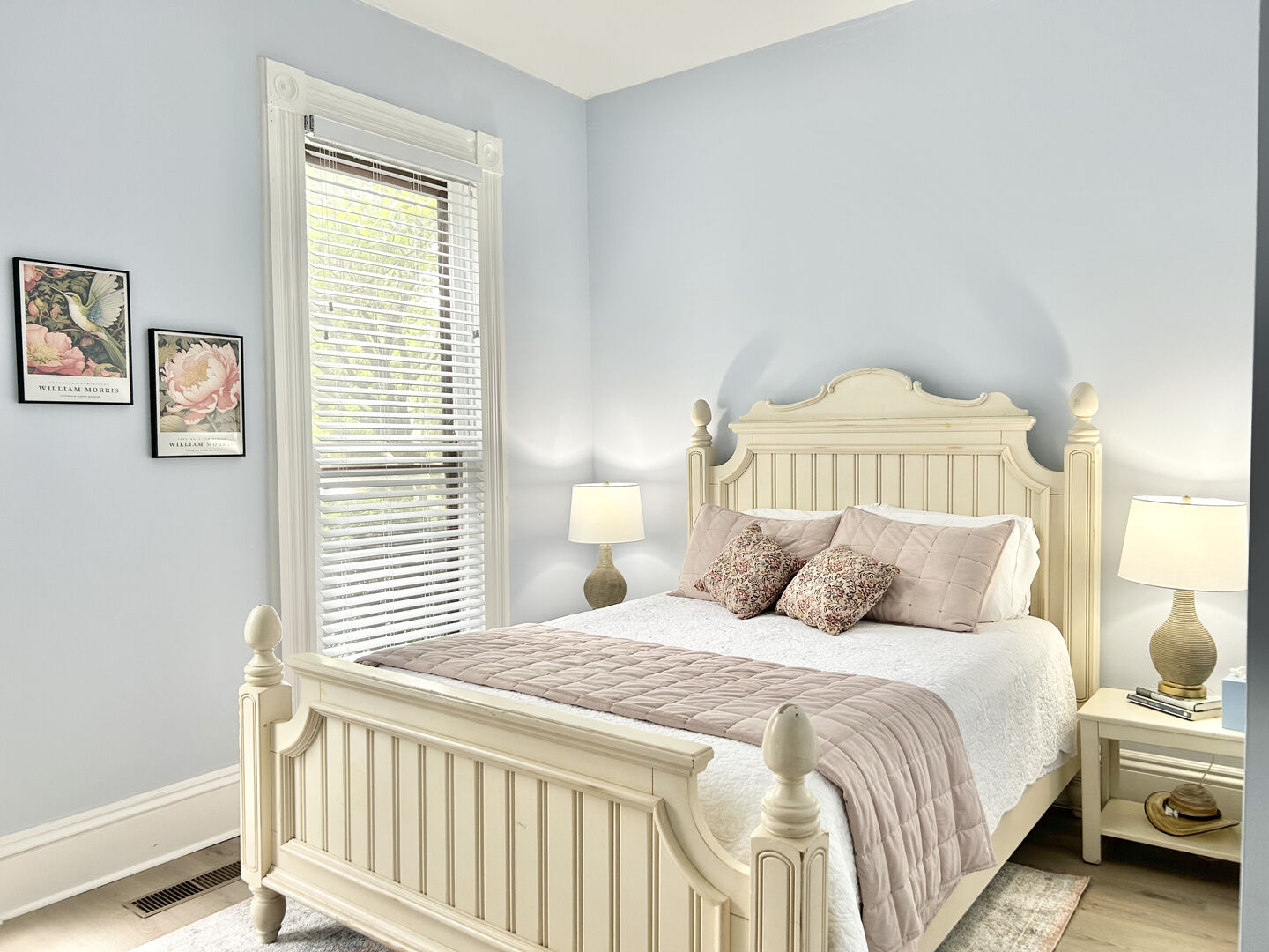 A serene bedroom features light blue walls, a cream wooden bed with a pink throw, two bedside tables with lamps, and framed art.