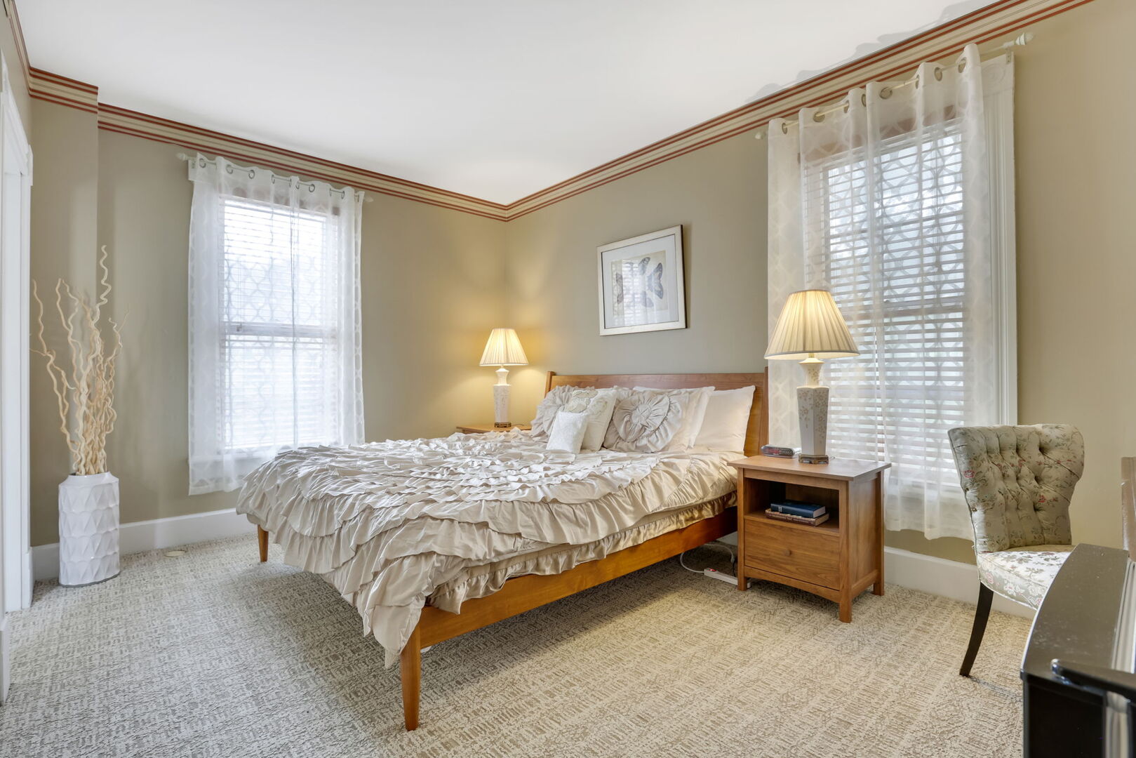 A neutral-toned bedroom with a wooden bed, ruffled bedding, two windows with sheer curtains, bedside tables with lamps, and decorative trim.