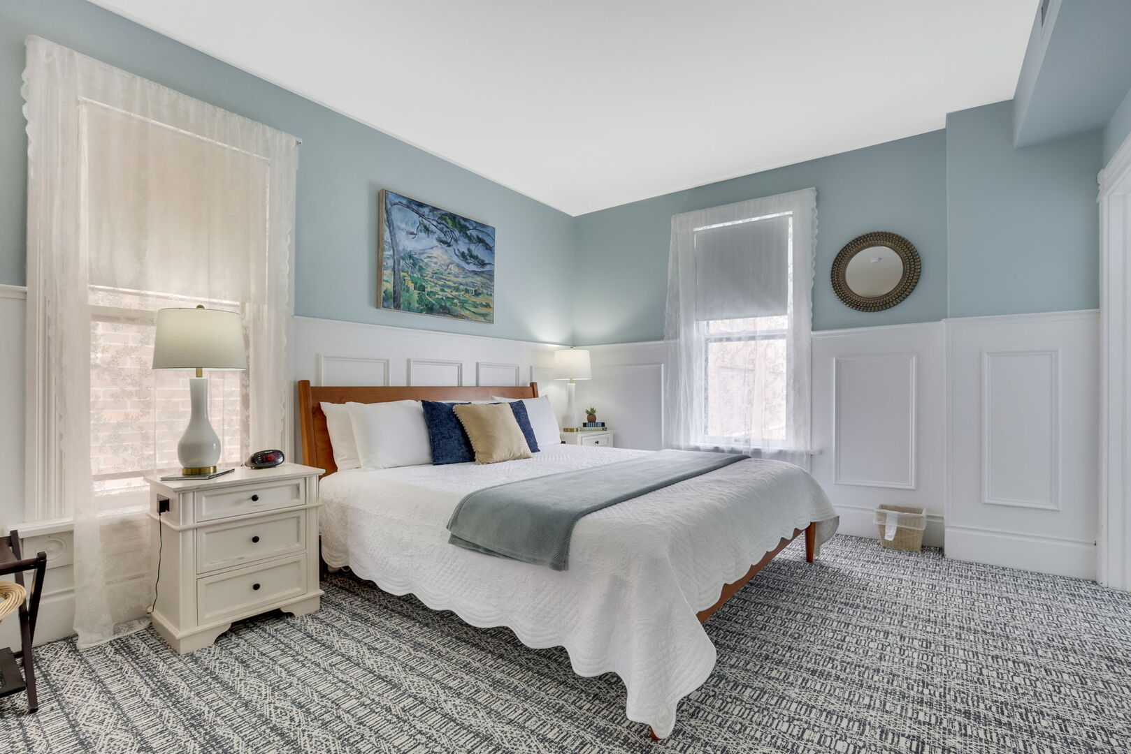 A light blue and white wainscoted bedroom with a wooden bed, two windows, a white bedside table with a lamp, and blue accent pillows.
