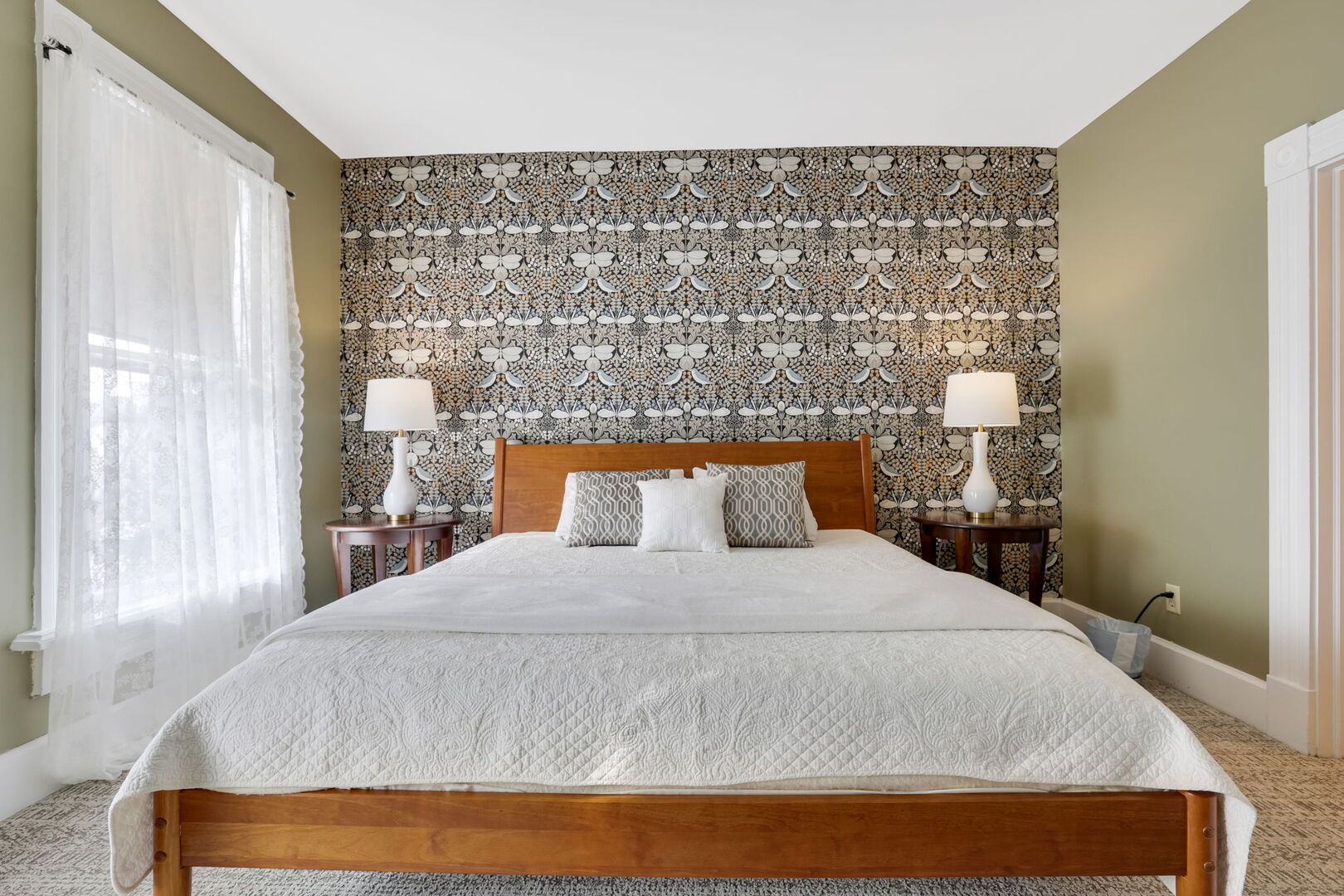 A chic bedroom features an accent wall with patterned wallpaper, olive green side walls, a wooden bed with white bedding, and two lamps.