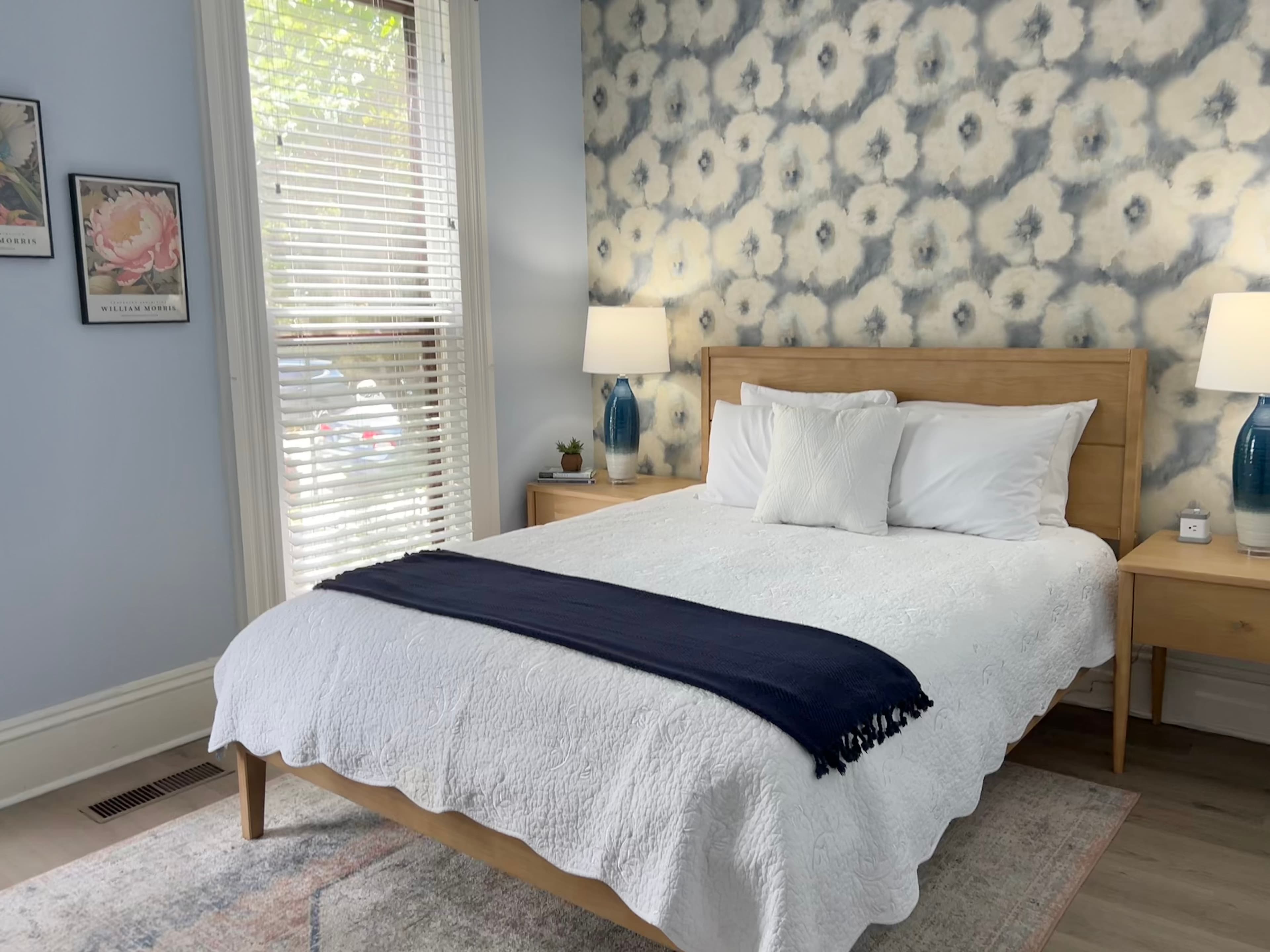 A serene bedroom features light blue walls, a light-oak wooden bed with a pink throw, two bedside tables with lamps, and framed art.