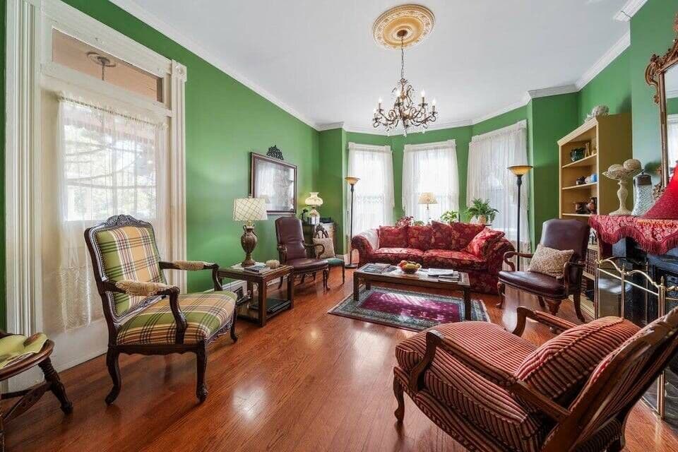A richly decorated living room with green walls, hardwood floors, a red sofa, patterned armchairs, and a ornate chandelier.