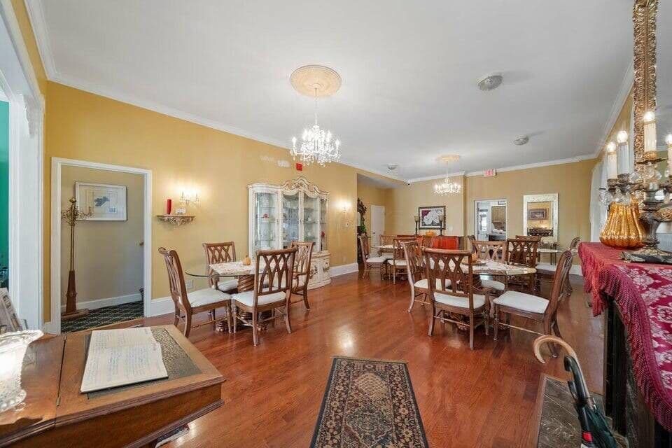 A spacious dining room with golden walls, hardwood floors, multiple tables and chairs, and elegant chandeliers, ready for guests.