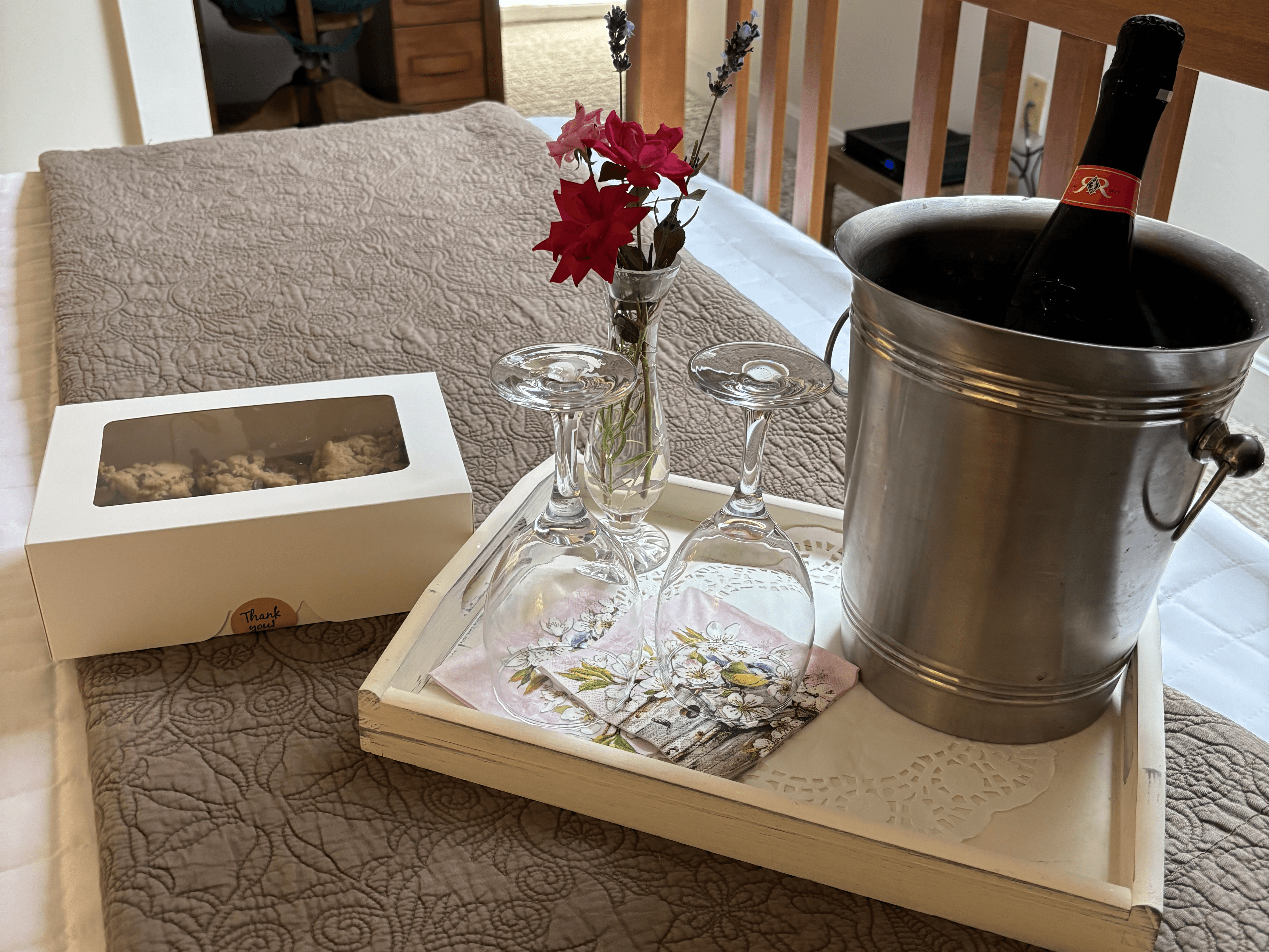 A tray with a silver ice bucket containing champagne, two glasses, a small vase of flowers, a decorative napkin, and a box of sweets, set on a bed.