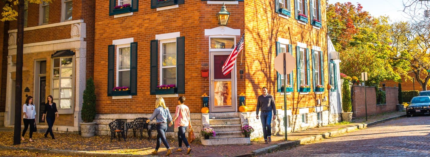 A sunny autumn day scene of a brick house with an American flag, featuring pedestrians walking along a tree-lined street.