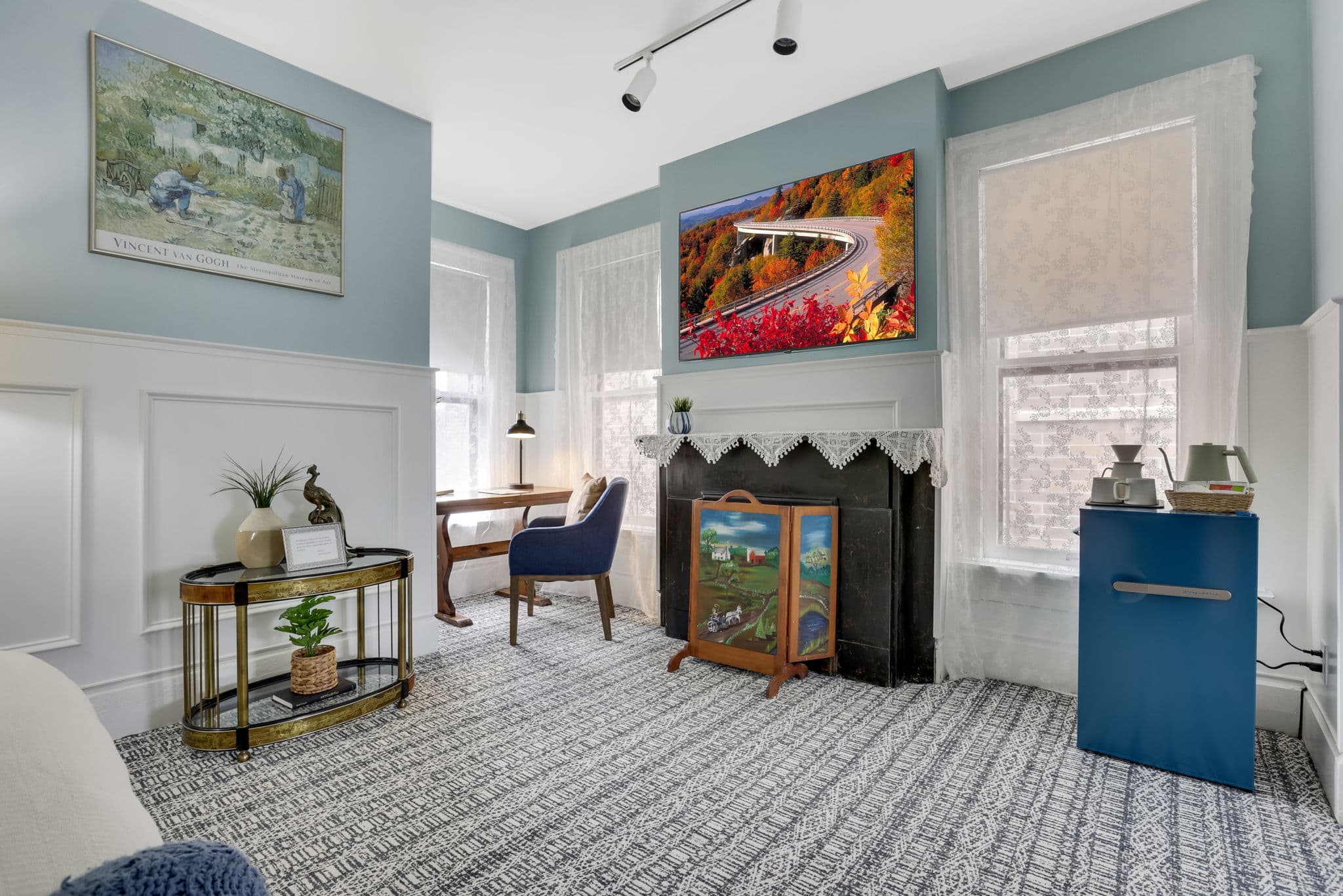 A cozy living room featuring a blue accent wall, a wall-mounted TV displaying autumn scenery, a wooden desk, and decorative artwork. A cozy living room featuring a blue accent wall, a wall-mounted TV displaying autumn scenery, a wooden desk, and decorative artwork.