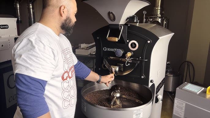 A man in a coffee shop operates a roasting machine, pouring freshly roasted coffee beans into a container. A man in a coffee shop operates a roasting machine, pouring freshly roasted coffee beans into a container.