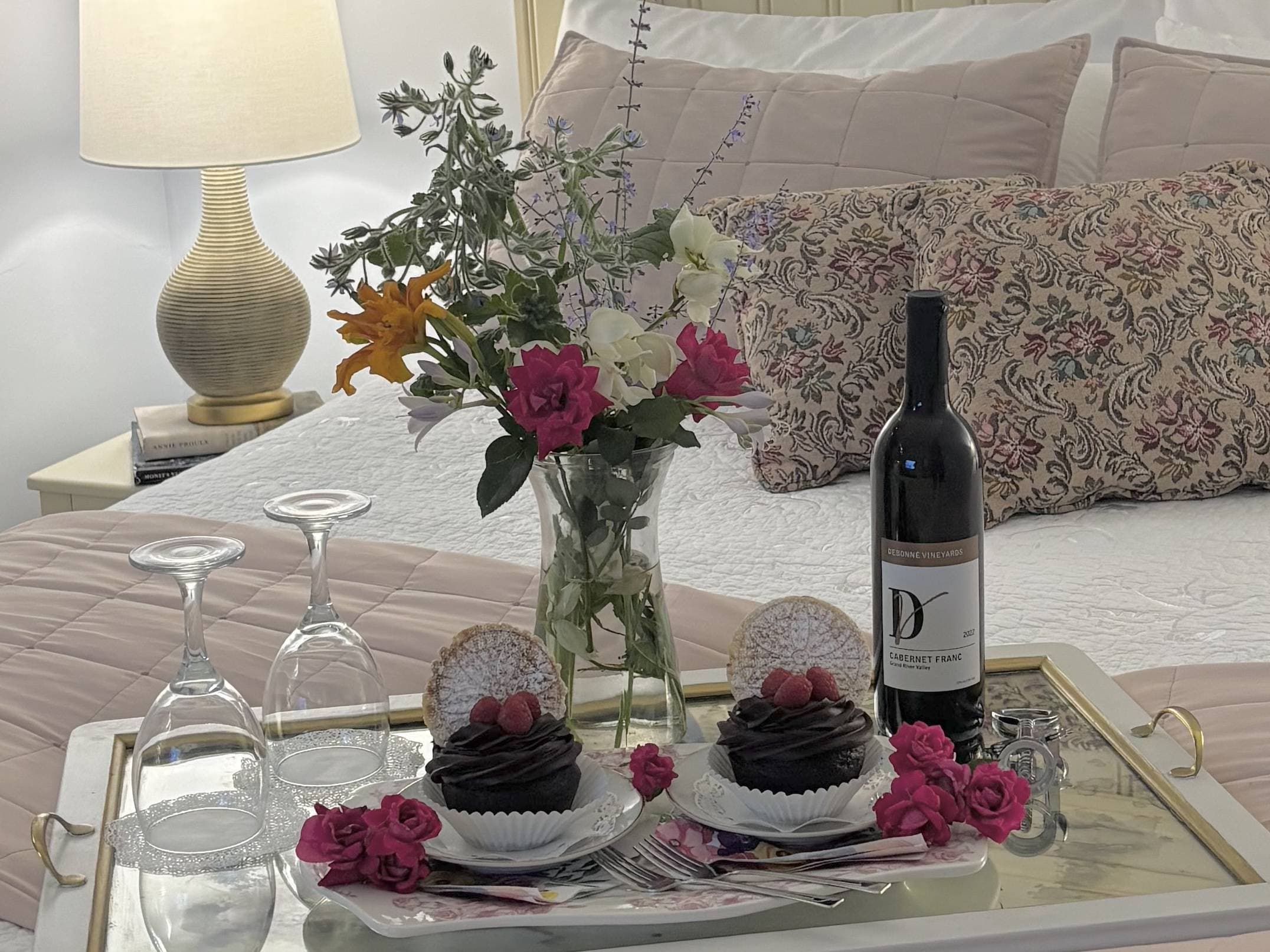 A cozy bedroom scene featuring a tray with chocolate cupcakes, a bottle of red wine, glasses, and a vase of mixed flowers beside a lamp.