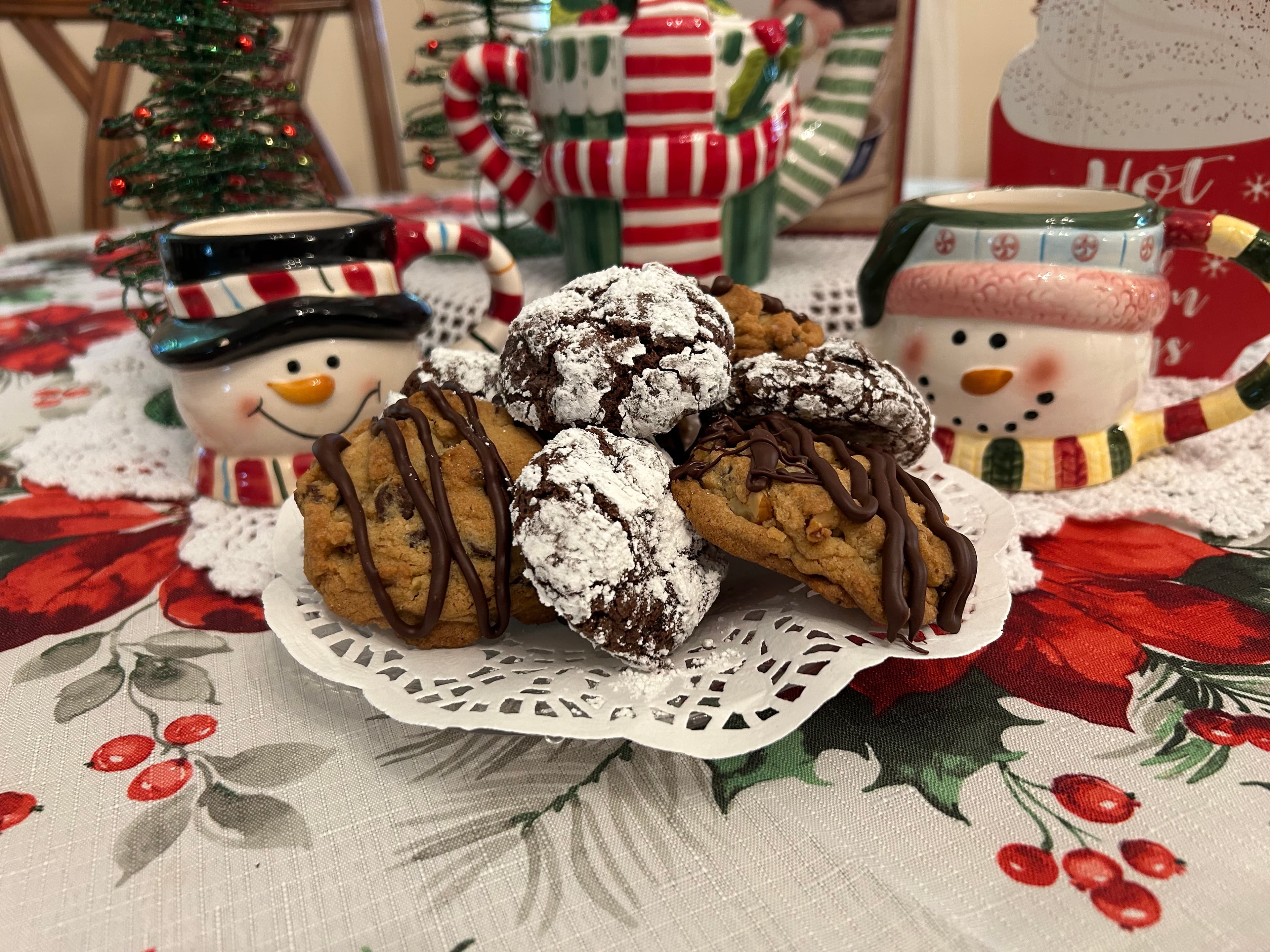 A plate of assorted holiday cookies sits on a festive table decorated with red and green patterns, alongside cheerful snowman mugs.