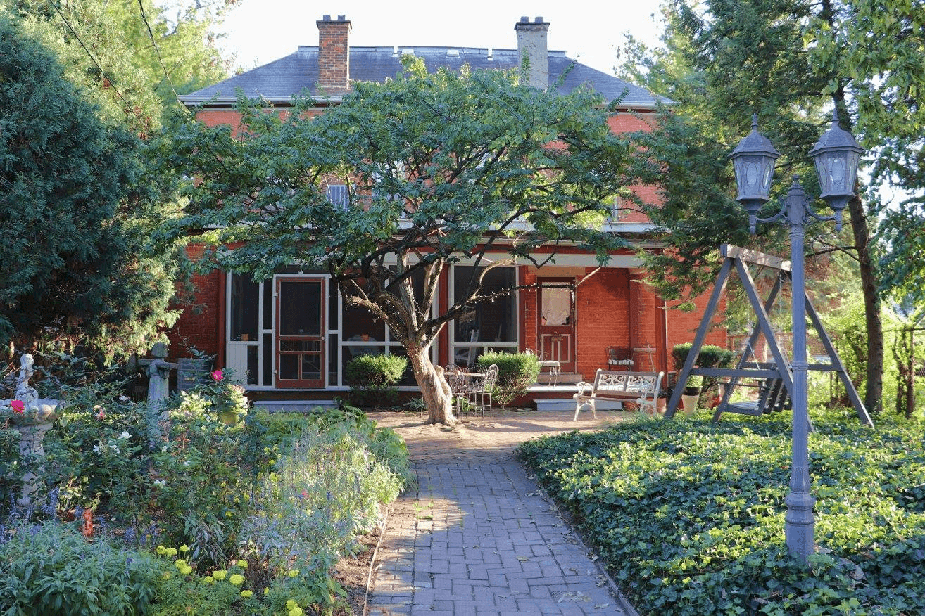Brick walkway surrounded by gardens on both sides, leading up to a tree directly in front of the back side of a large brick two story house