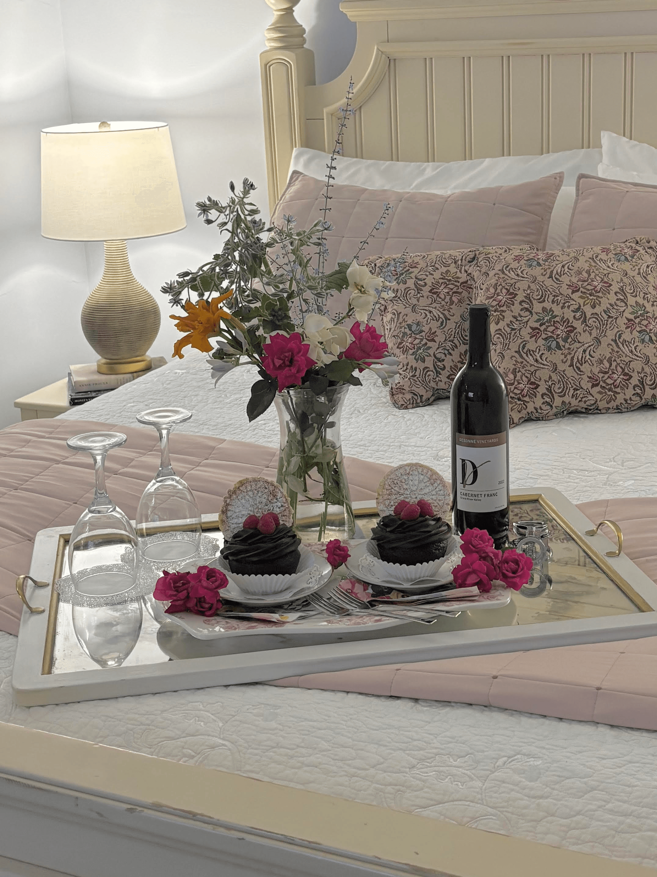 A beautifully set tray on a bed featuring two desserts, a bottle of wine, glasses, and a vibrant floral arrangement.
