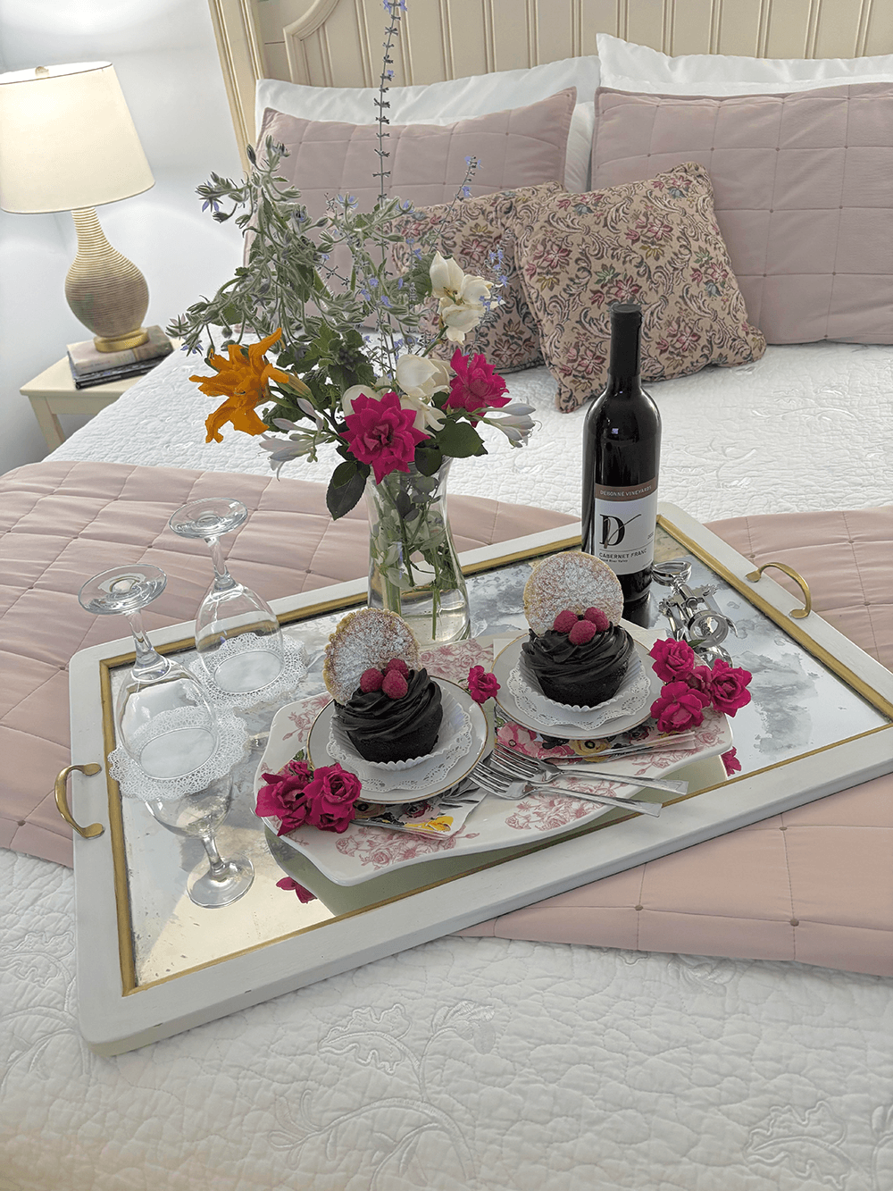 A beautifully set tray on a bed featuring two desserts, a bottle of wine, glasses, and a vibrant floral arrangement.