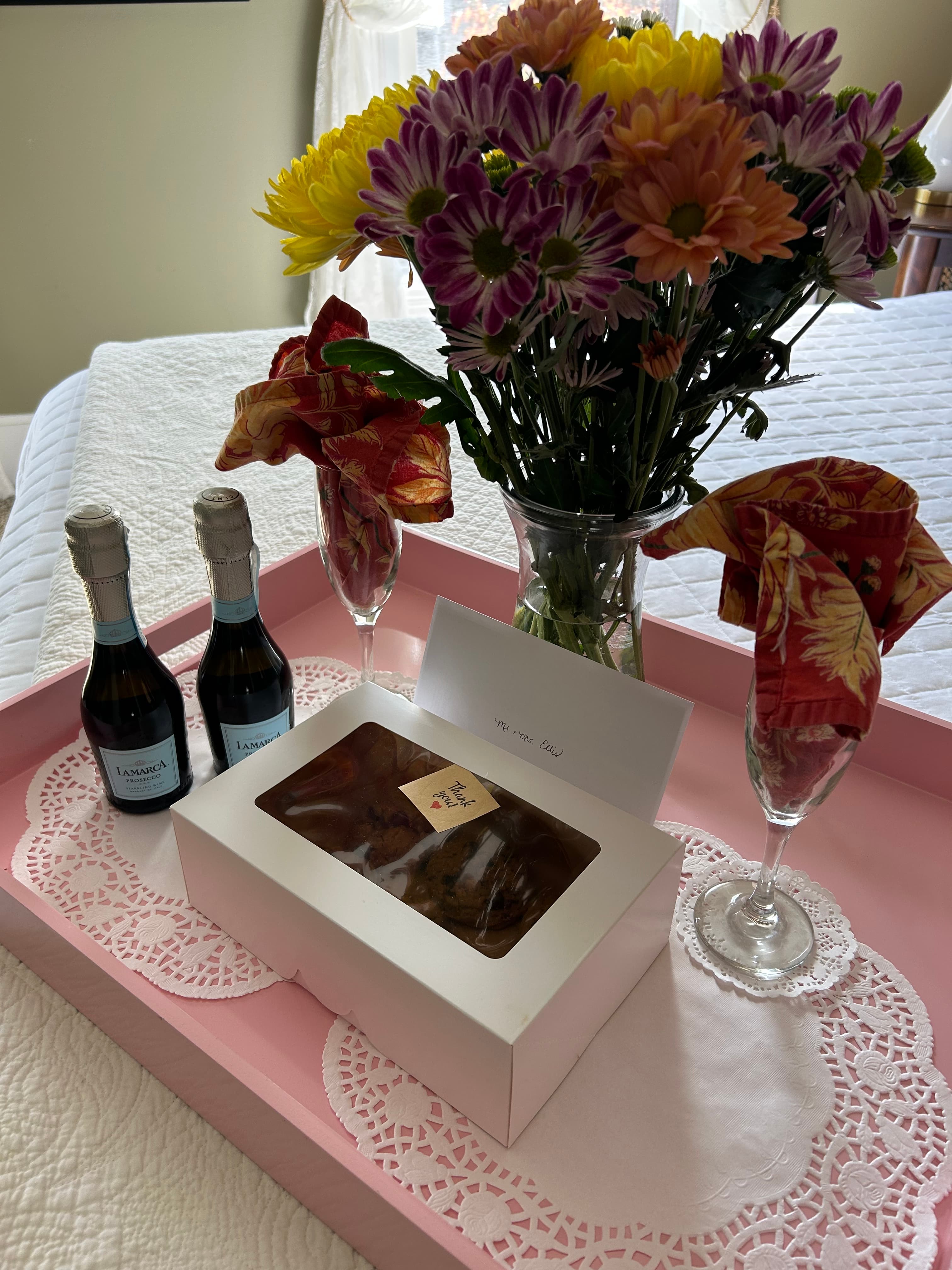 A pink tray holds two mini sparkling bottles, a box of treats, a card, and a bouquet of colorful flowers.