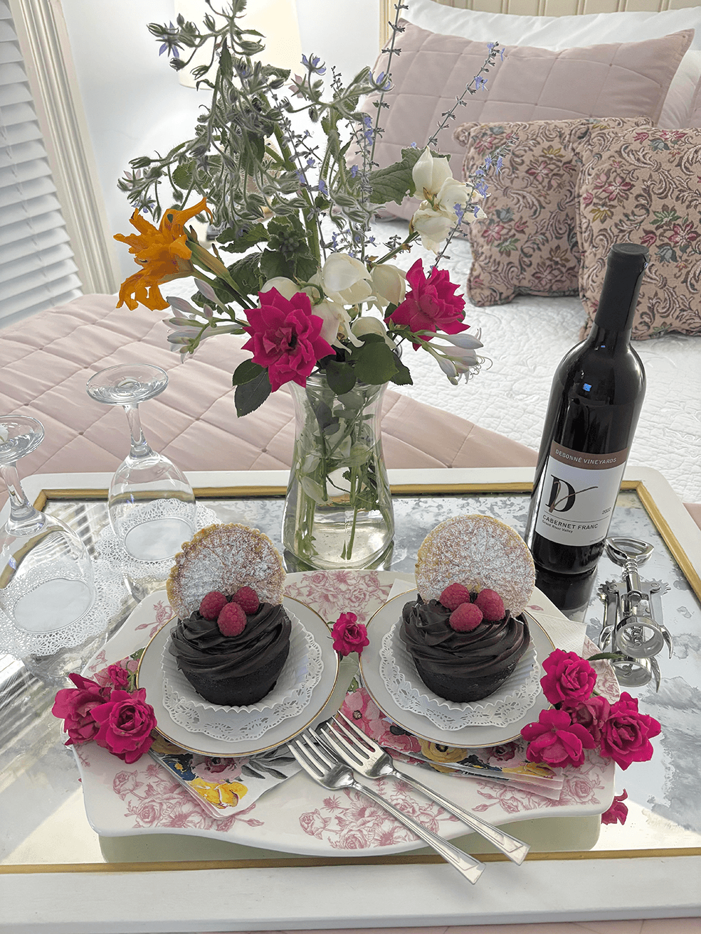 A beautifully arranged table features two chocolate cupcakes with raspberries, a bouquet of flowers, wine, and elegant glassware.